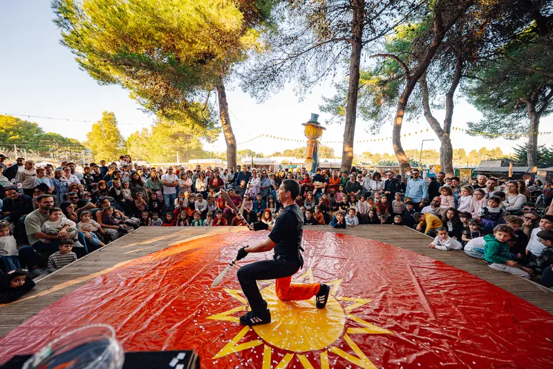 Il giocoliere sulla pista rossa del circo, circondato dal pubblico seduto fra i pini — Parco d’Autunno 2026 al Raffo Parco Gondar di Gallipoli